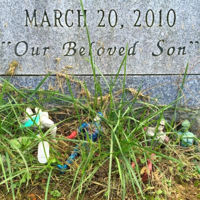 four plastic action figures in weeds in front of gravestone with date and epitaph