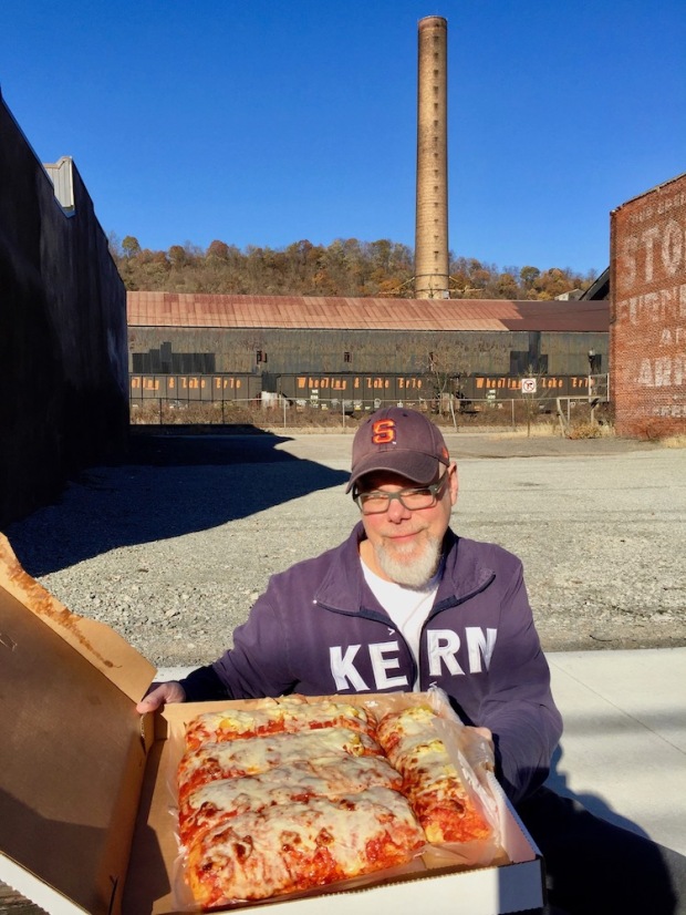 man holding open box of pizza with smoke stack and steel mill behind him