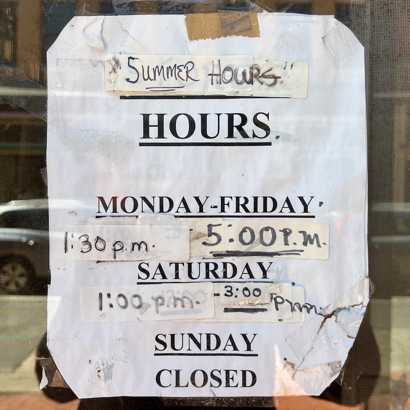 store hours posted in shop window with many hand alterations